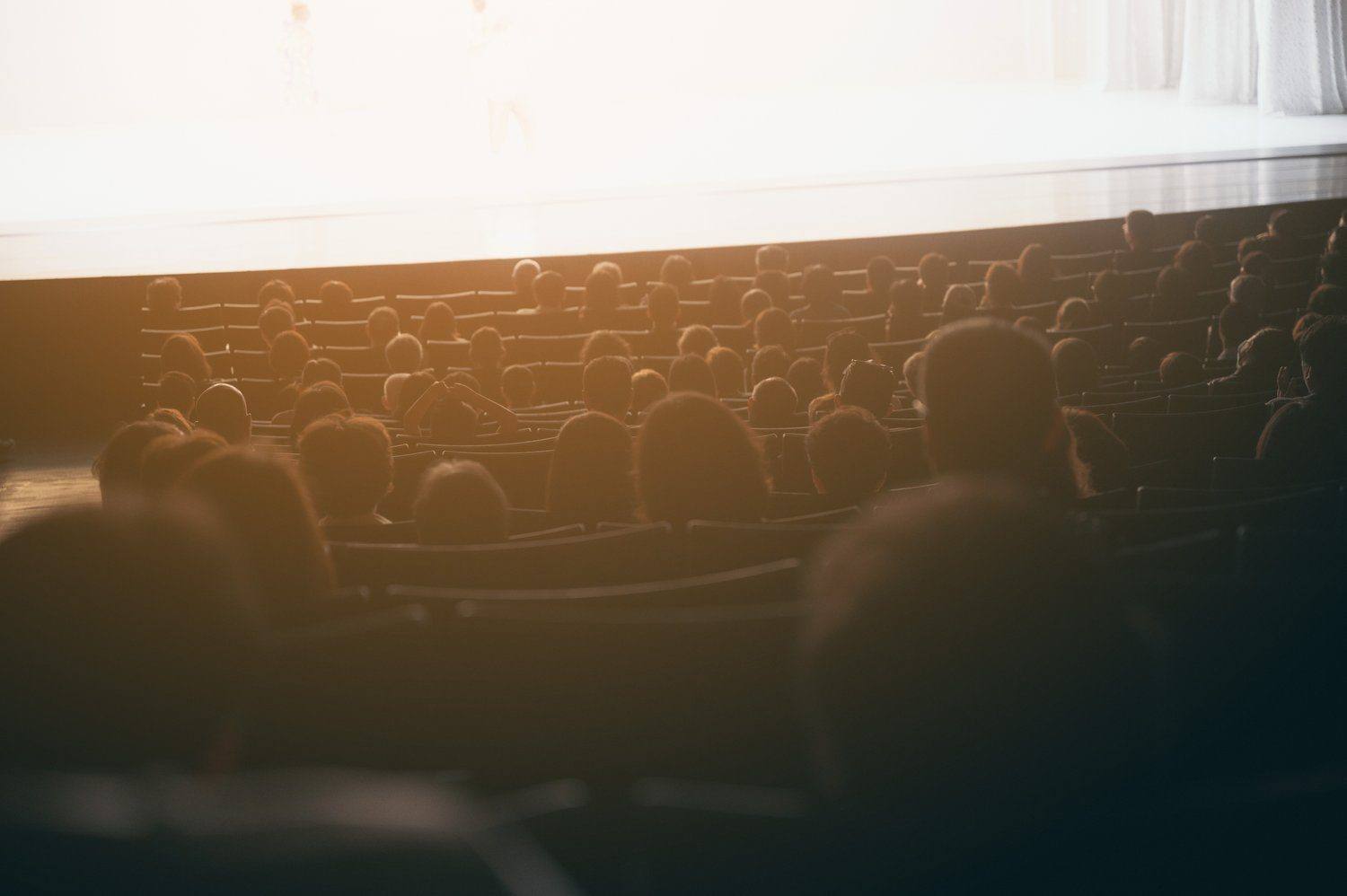 Widzowie od tyłu w kameralnej sali teatralnej, przed nimi przyciemniona scena