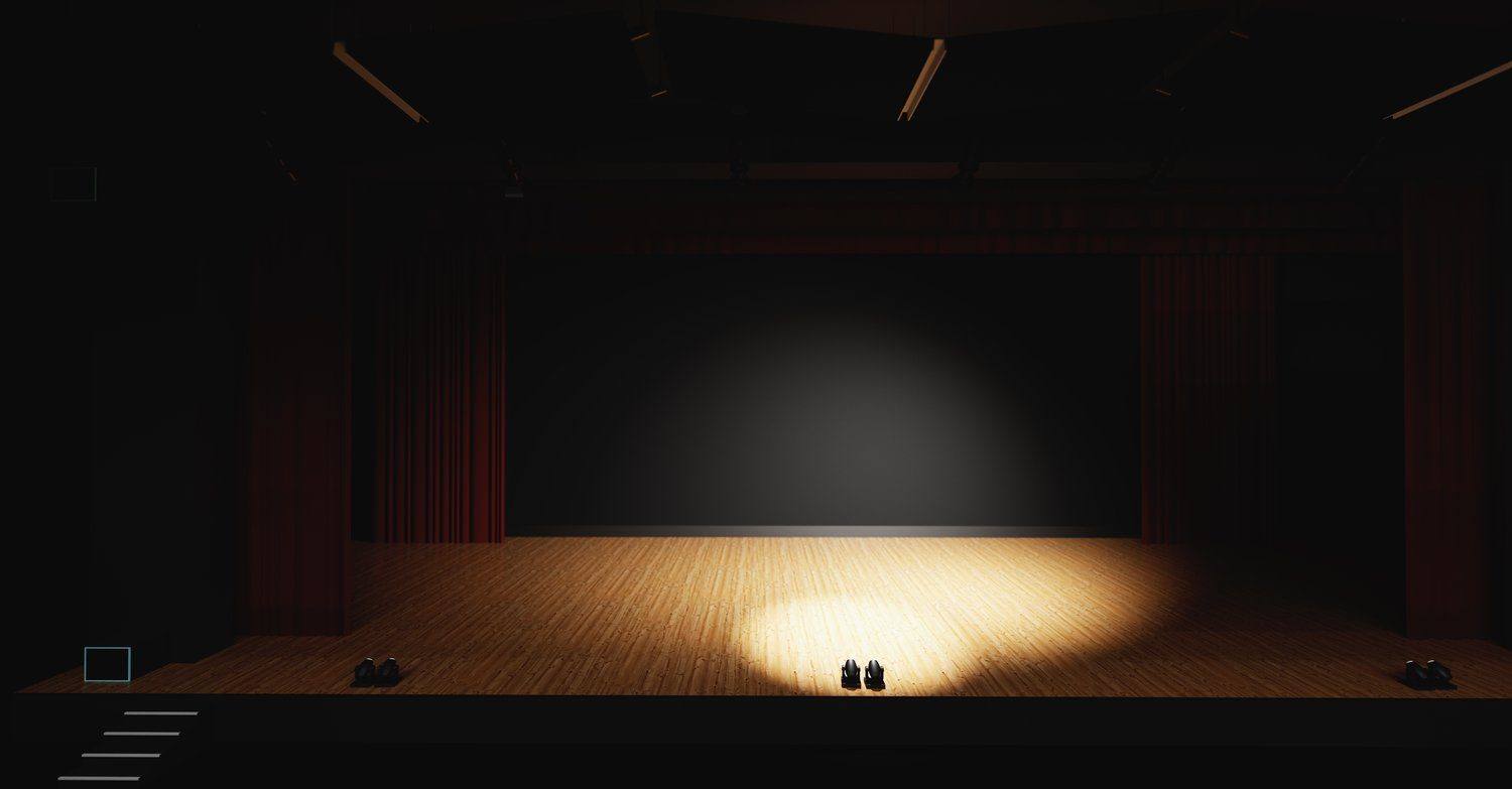 Niewielka czarna scena teatralna oświetlona ciepłym punktowym światłem, przed nią puste krzesła